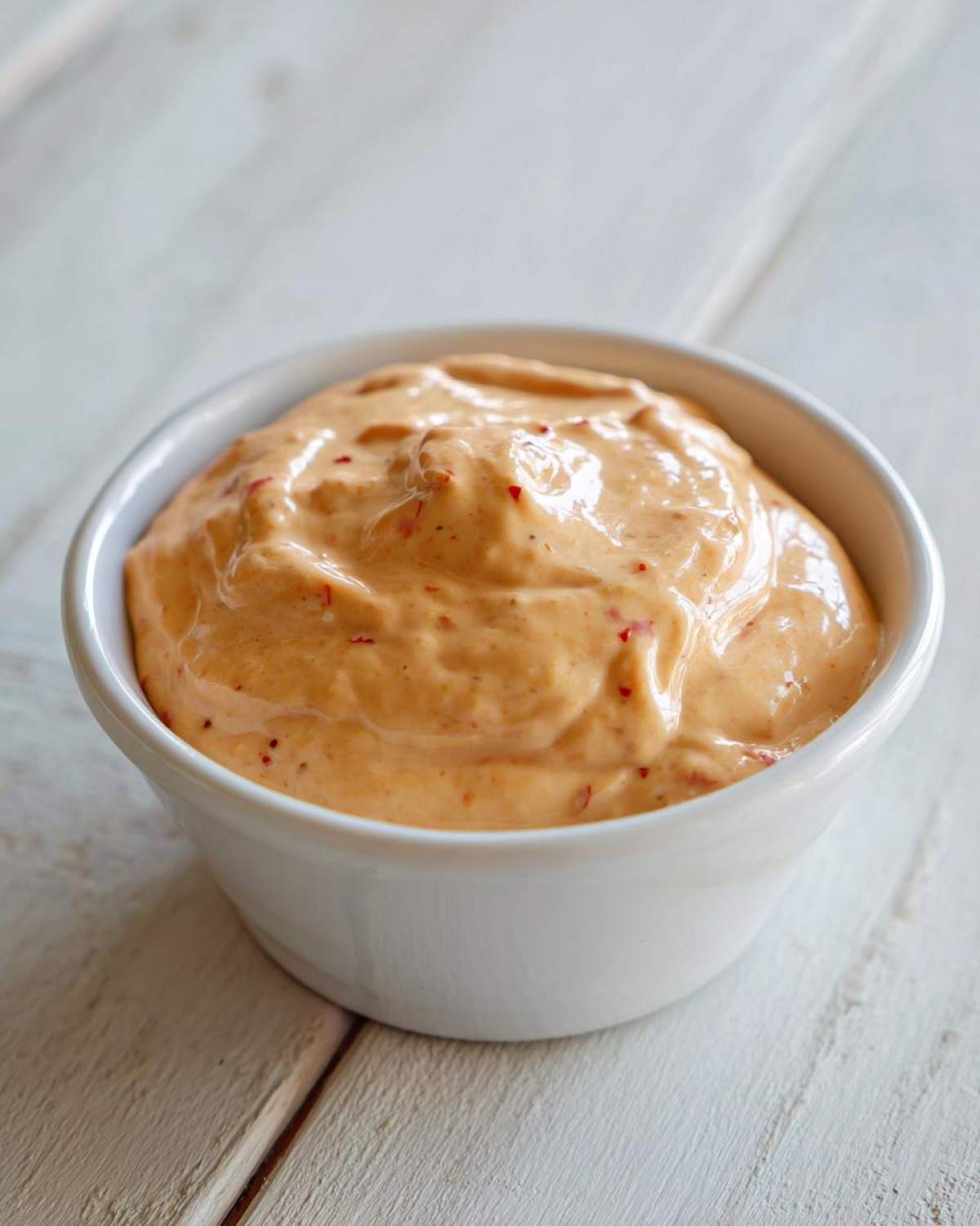 Close-up of creamy, orange-hued burger sauce with visible red spice flecks in a small white bowl.