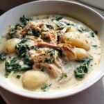 Close-up of a bowl of creamy chicken and gnocchi soup with wilted spinach and herbs.