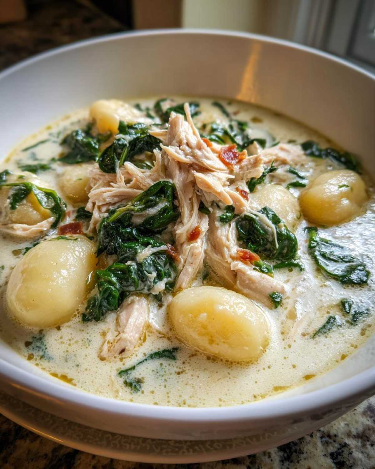 Close-up of a hearty bowl of creamy gnocchi soup topped with shredded chicken, wilted spinach, and bacon bits.