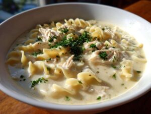 Close-up of a bowl of creamy chicken soup featuring shredded chicken, wide egg noodles, and fresh parsley garnish.