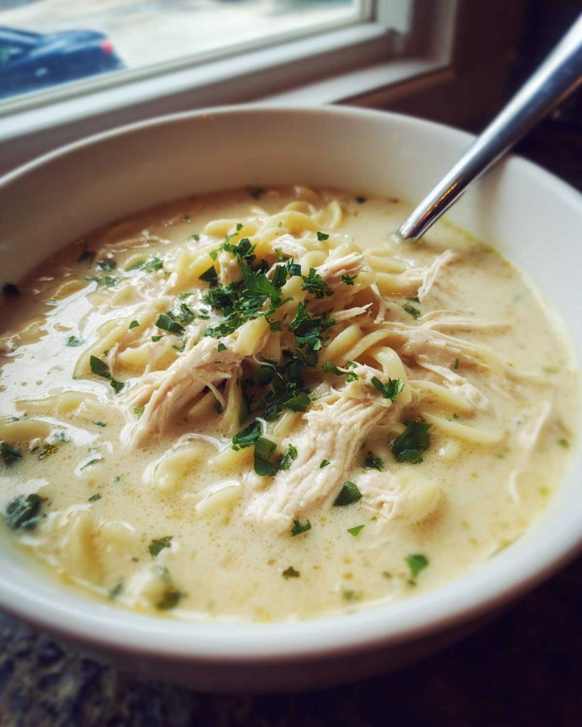 Close-up of a bowl of creamy chicken soup with egg noodles, shredded chicken, and fresh parsley.