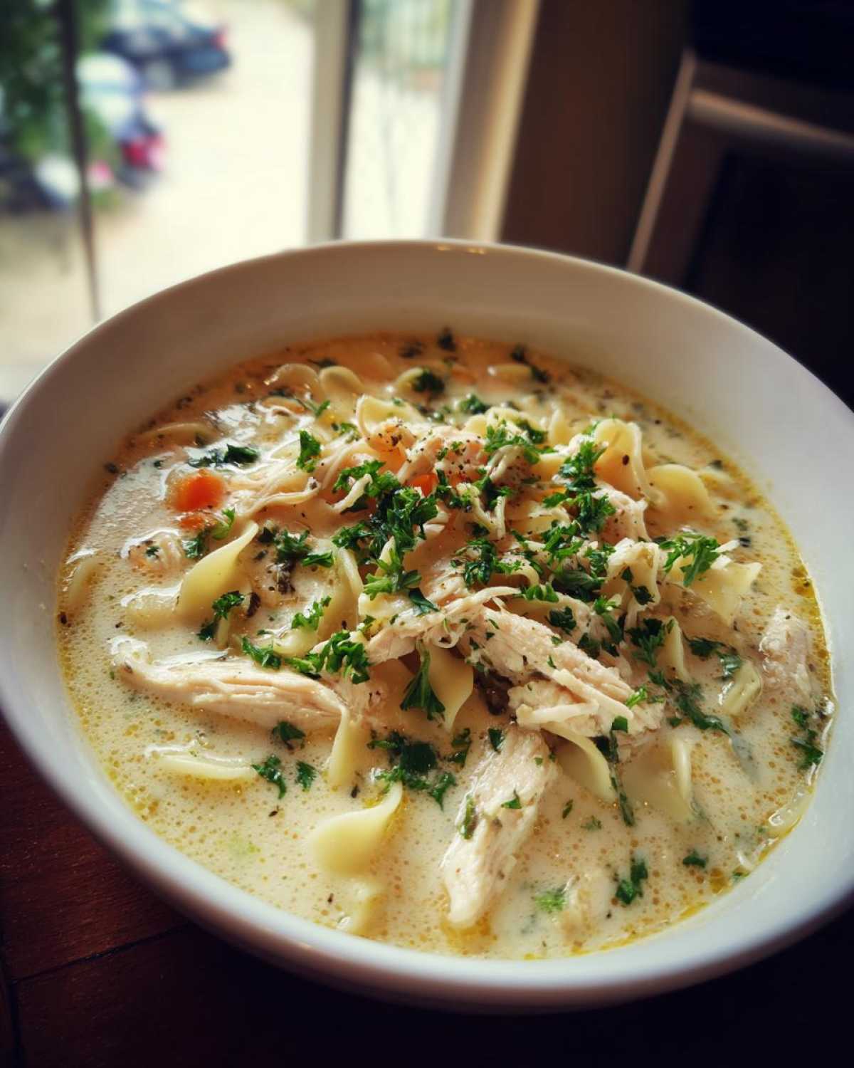 A close-up of a bowl of creamy chicken soup featuring shredded chicken, egg noodles, and fresh parsley.