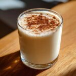 A glass of creamy, light-colored coquito recipe topped generously with ground cinnamon.