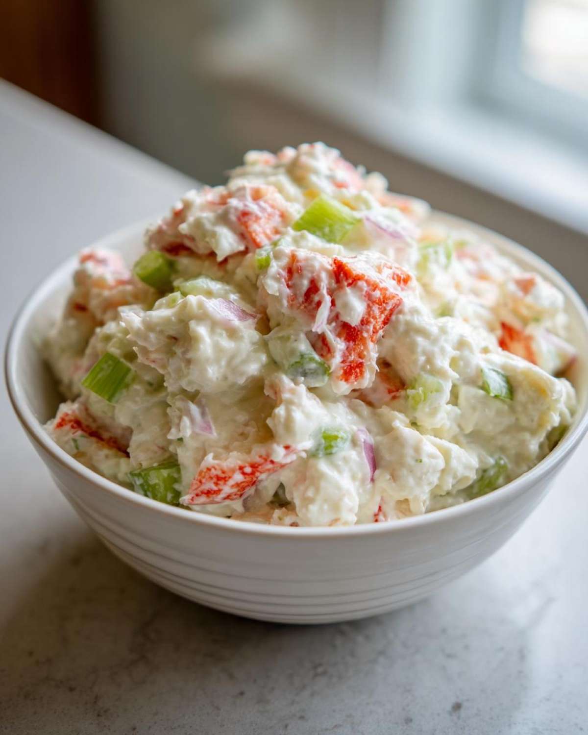 A close-up of a white bowl filled with creamy crab salad featuring chunks of red imitation crab, green celery, and red onion.