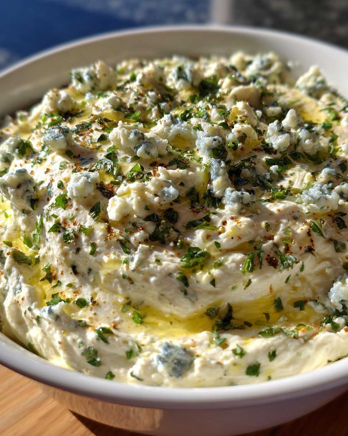 Close-up of creamy dirty martini dip topped with crumbles of blue cheese, chopped parsley, and olive oil drizzle.