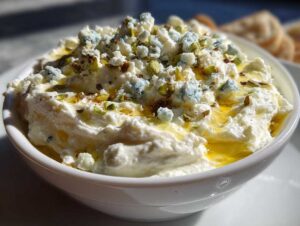 Close-up of a creamy white dirty martini dip topped with crumbles of blue cheese, chopped pistachios, and olive oil.