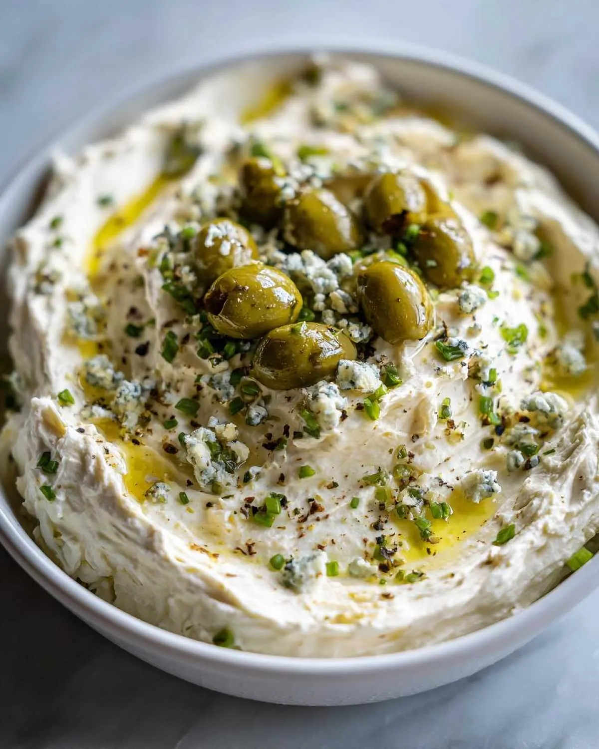 Close-up of a creamy white dirty martini dip garnished with green olives, blue cheese crumbles, and chives.