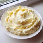 A white bowl filled with incredibly creamy garlic mashed potatoes, showing swirls and a pat of melting butter on top.