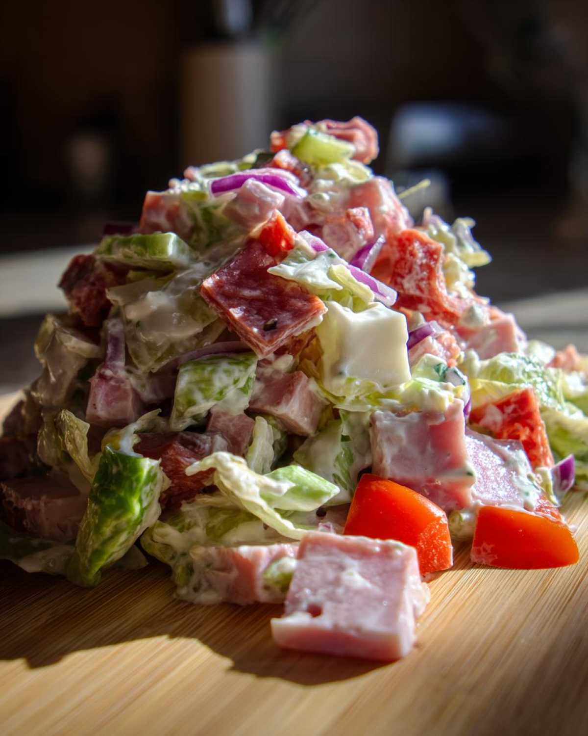 A mound of creamy grinder salad featuring diced ham, salami, cheese, lettuce, tomato, and red onion on a wooden board.