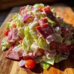 A vibrant, creamy grinder salad piled high with chopped lettuce, salami, ham, cheese cubes, red onion, and cherry tomatoes.