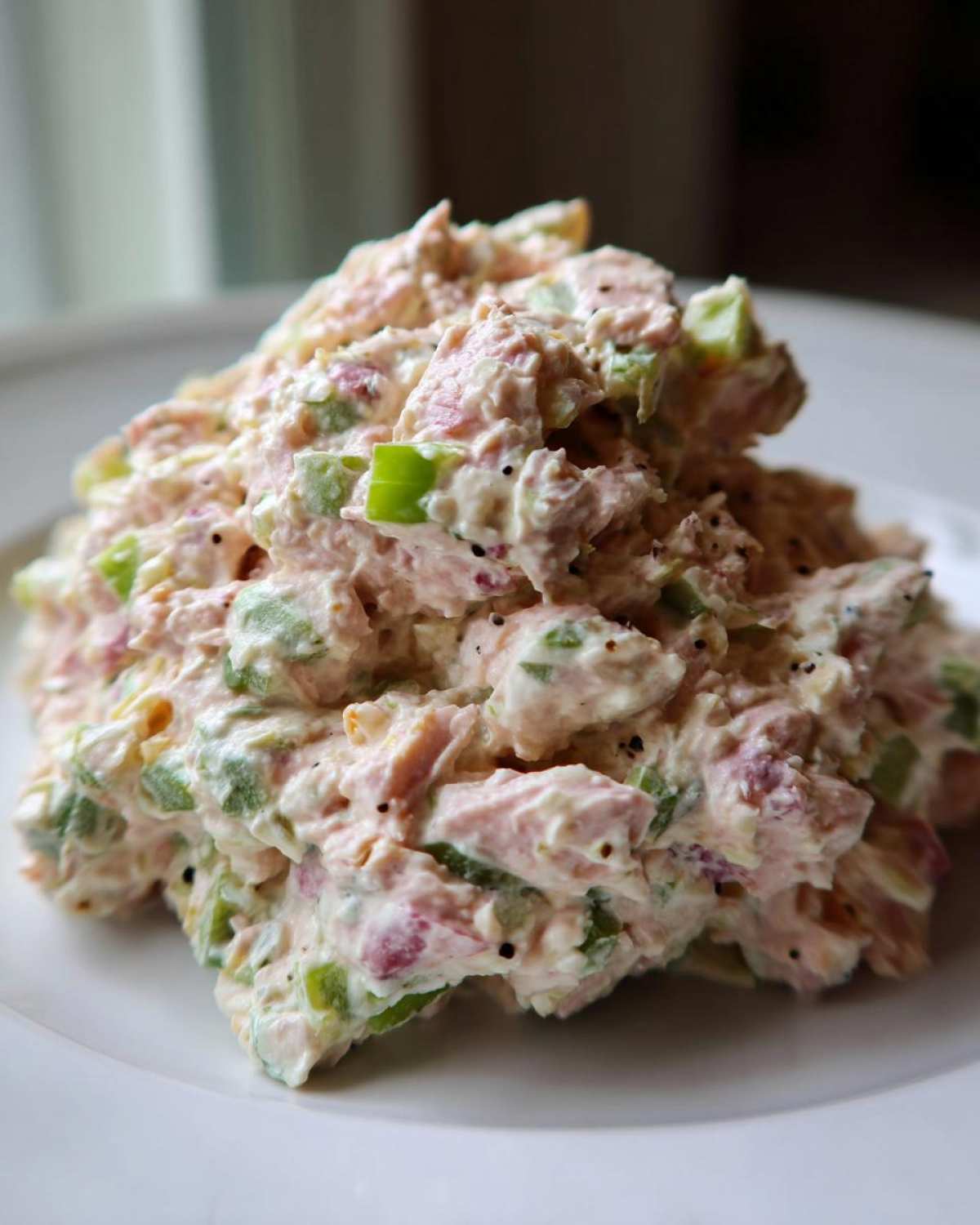 A mound of creamy, pink ham salad mixed with visible chunks of green celery and black pepper, served on a white plate.