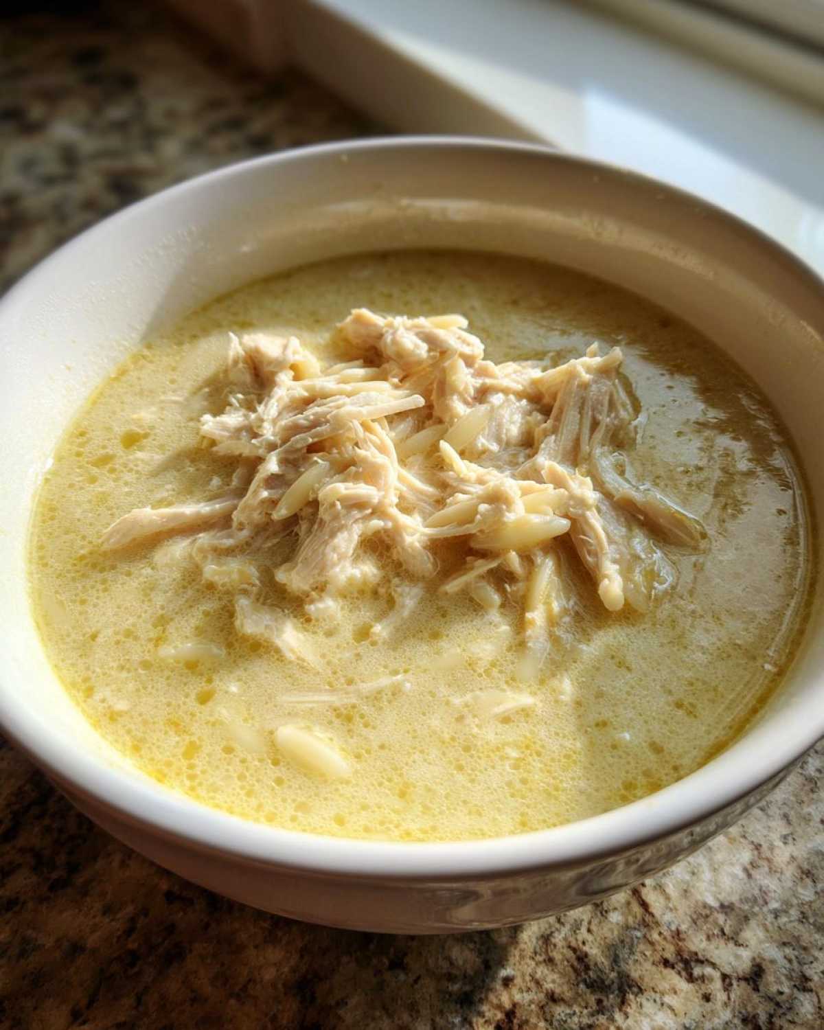 A close-up of a bowl of creamy lemon chicken soup topped generously with shredded chicken and orzo pasta.