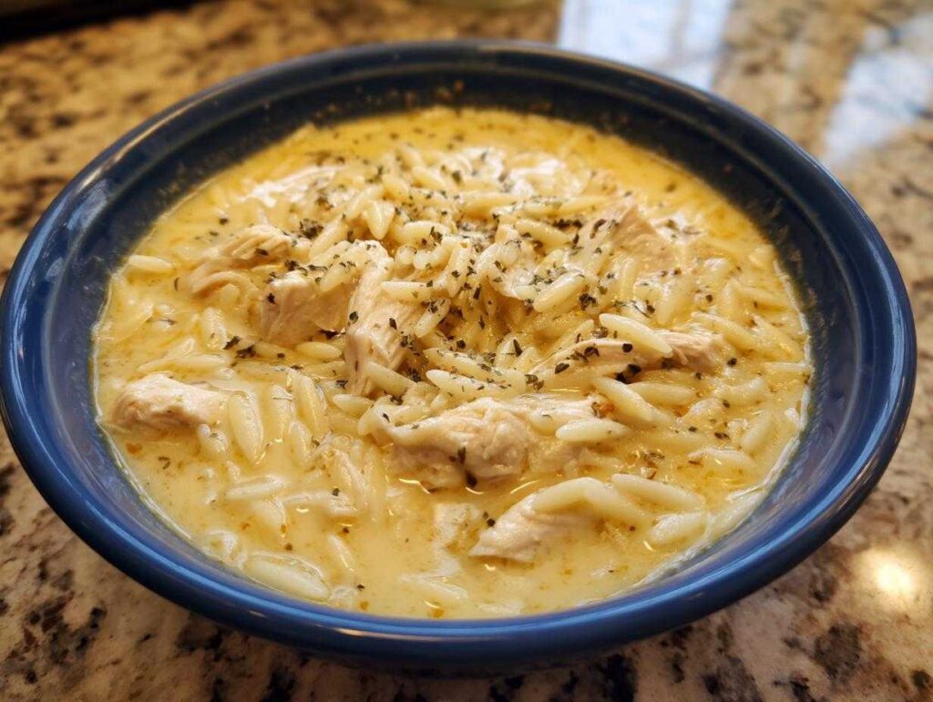 Close-up of creamy lemon chicken soup featuring chunks of chicken and orzo pasta, garnished with dried herbs.
