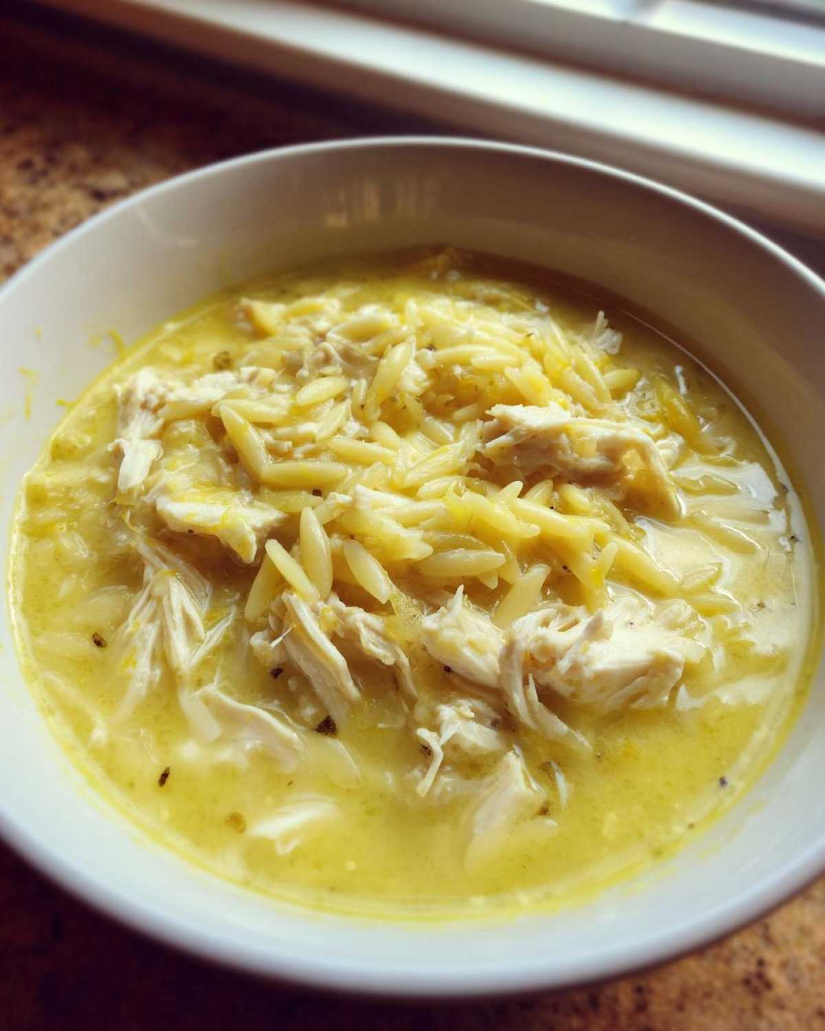 Close-up of a white bowl filled with creamy lemon chicken soup, featuring shredded chicken and orzo pasta.