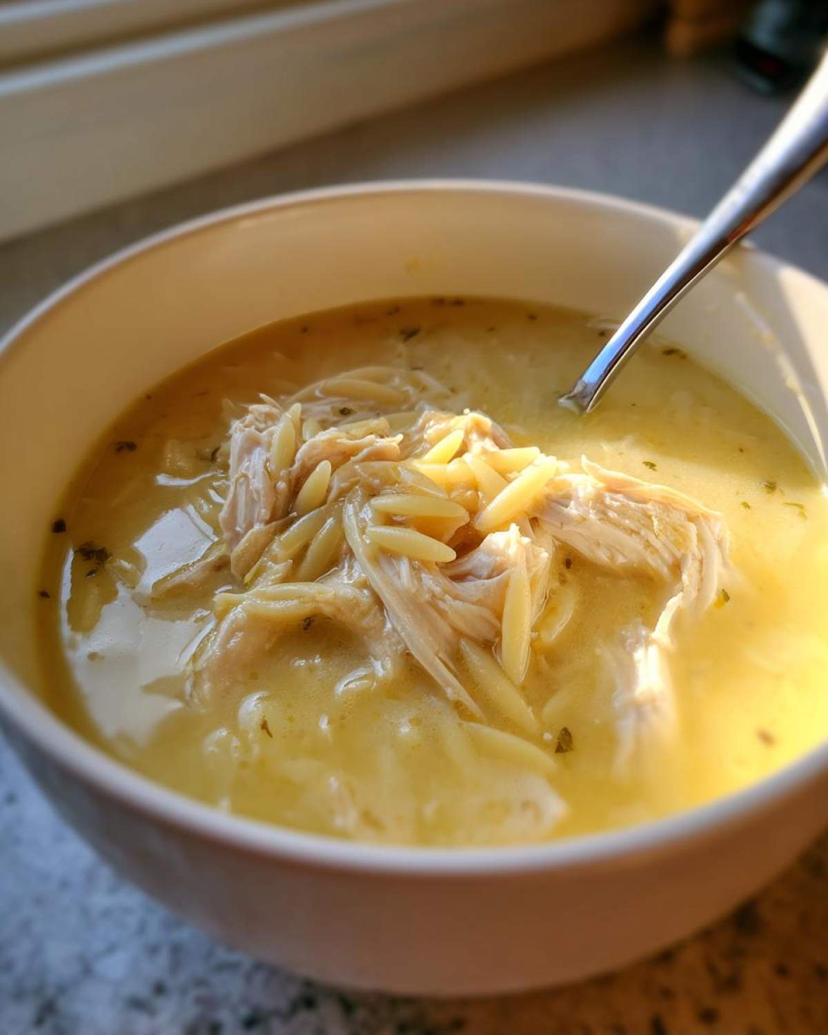 A close-up of creamy lemon chicken soup featuring shredded chicken and orzo pasta in a white bowl.