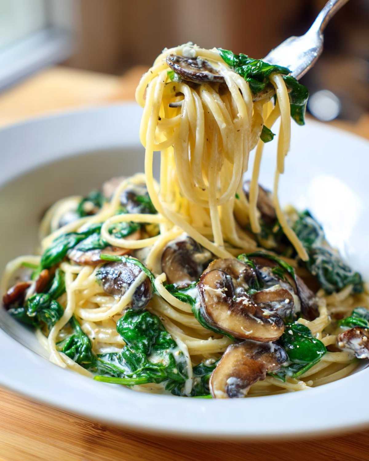 A fork lifting a twirl of spaghetti from a bowl of creamy mushroom pasta mixed with wilted spinach.
