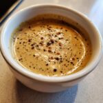 A close-up of creamy peppercorn sauce topped generously with cracked black and red peppercorns in a white bowl.
