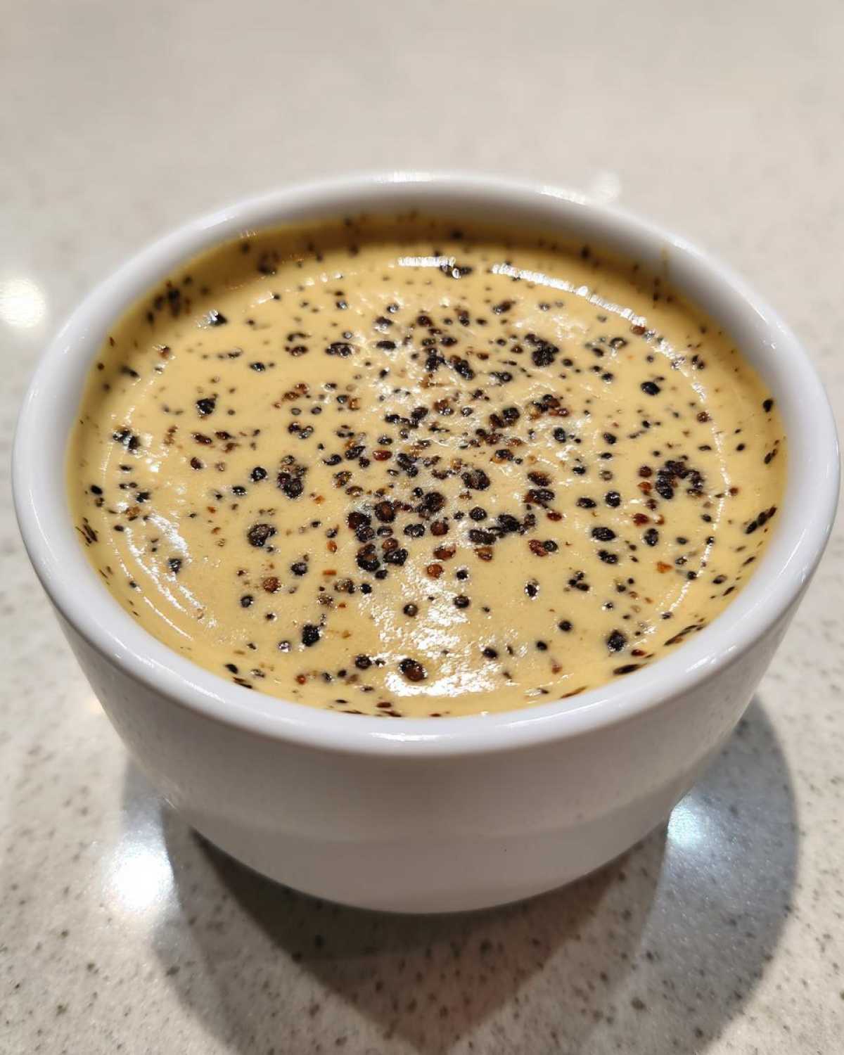Close-up of a rich, creamy peppercorn sauce topped generously with cracked black peppercorns in a small white bowl.