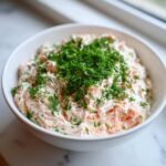 A white bowl filled with creamy salmon dip, mixed with pink salmon pieces and heavily garnished with fresh green dill.