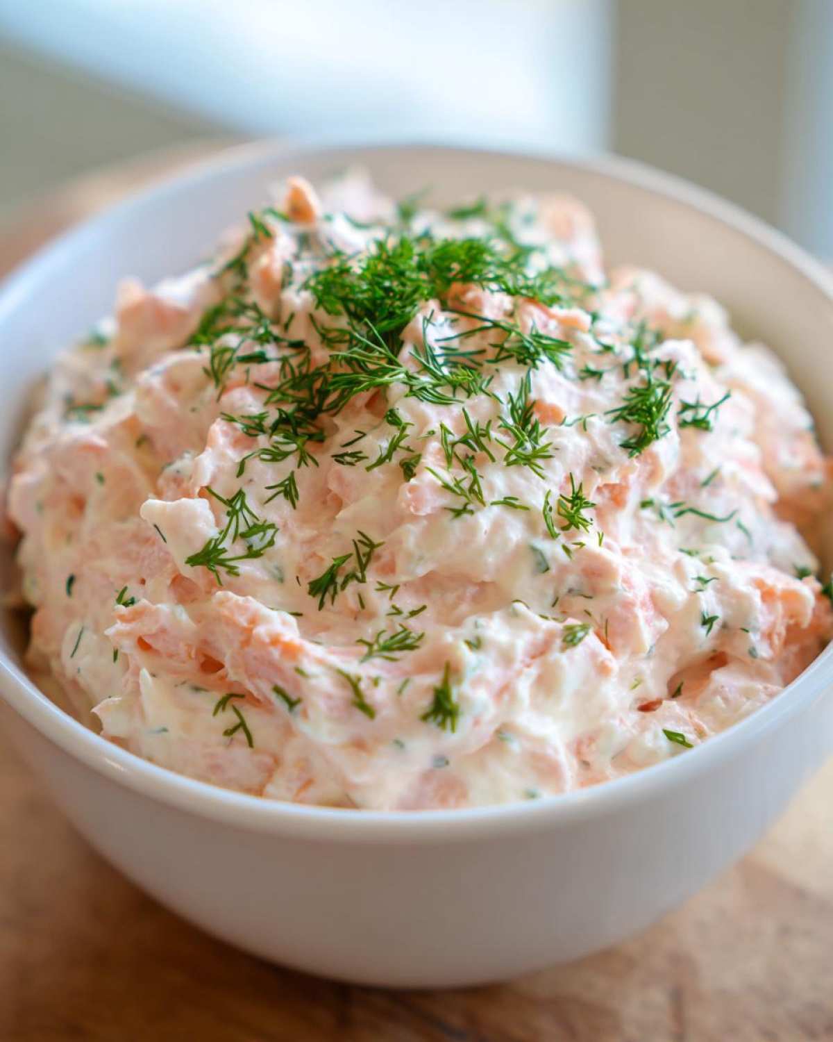 Close-up of a bowl filled with creamy salmon dip, featuring pink salmon chunks mixed with a white base and garnished heavily with fresh green dill.