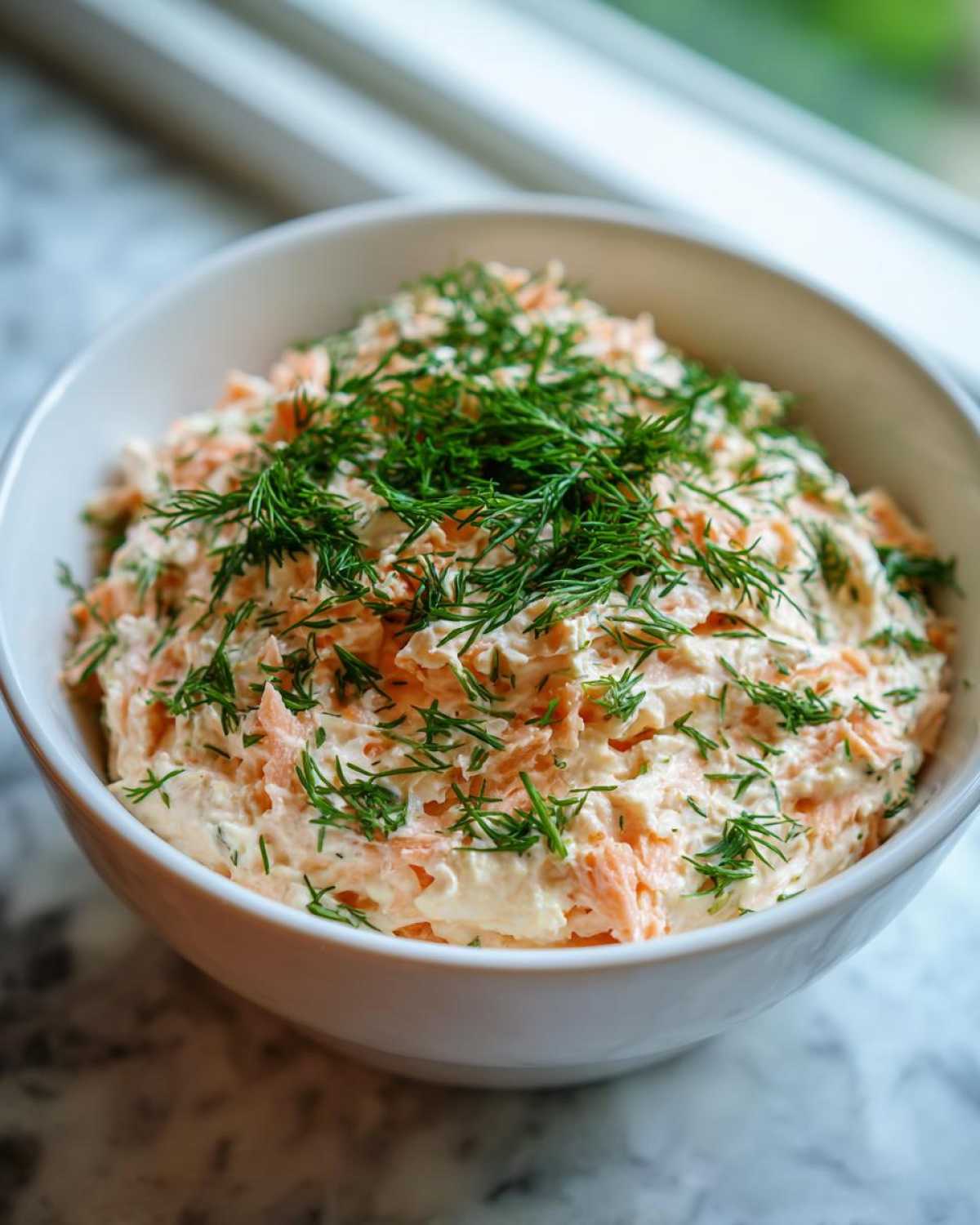 A white bowl filled with creamy salmon dip, generously topped with fresh chopped dill.