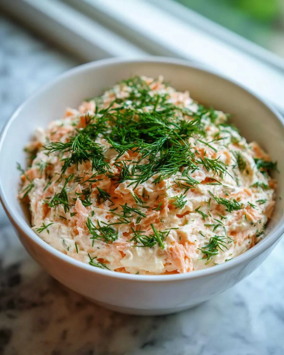A white bowl filled with creamy salmon dip, generously topped with fresh chopped dill.
