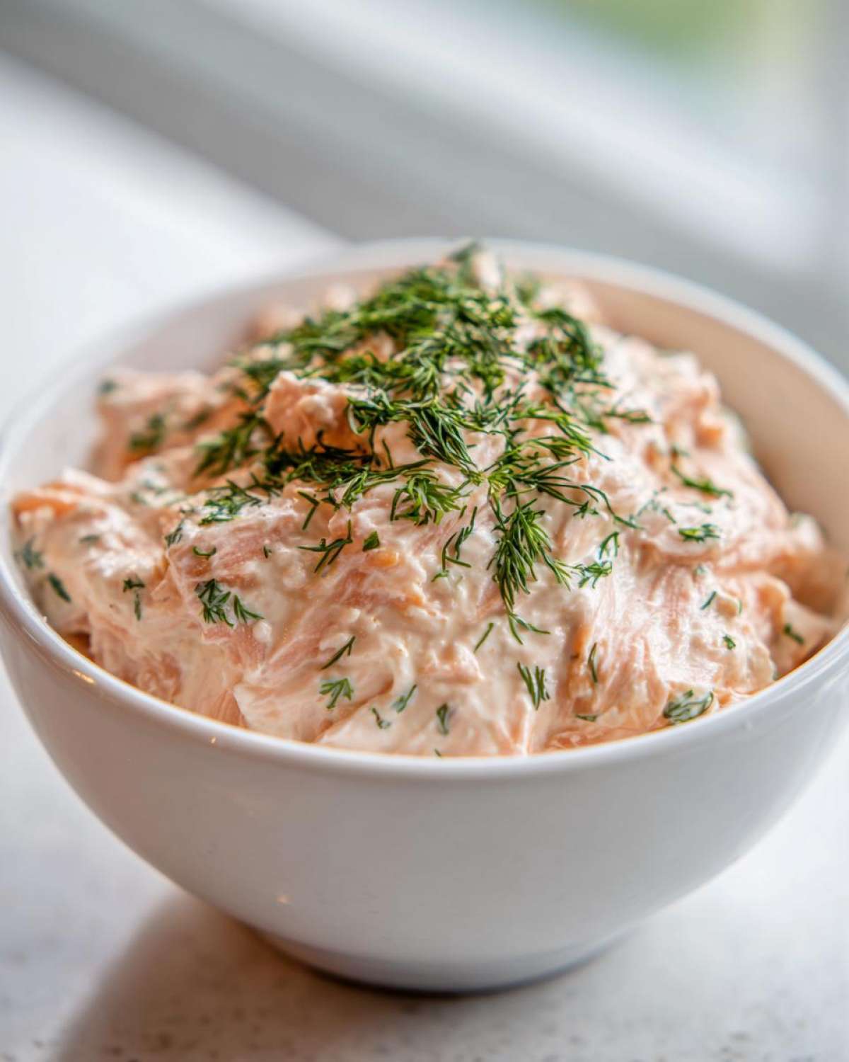 A close-up of creamy salmon dip mixed with herbs, generously topped with fresh dill in a white bowl.