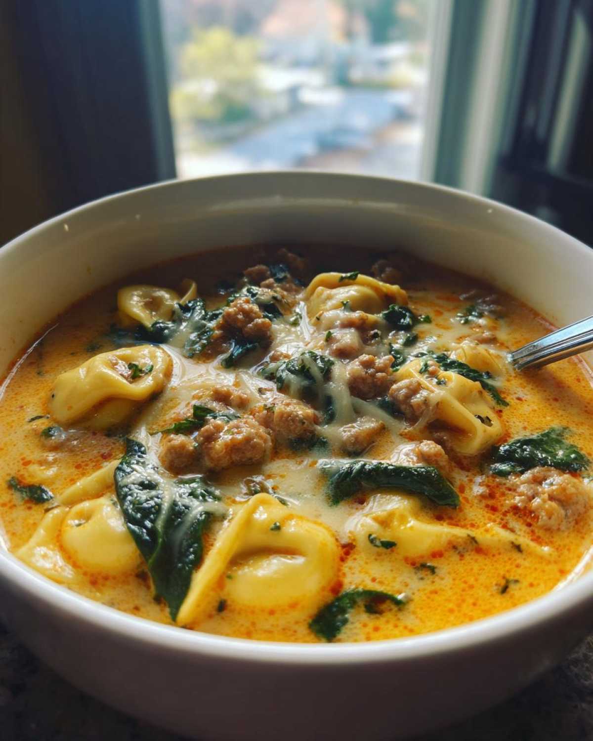 Close-up of a hearty bowl of creamy sausage tortellini soup with spinach and melted cheese.