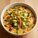 A white bowl filled with creamy, orange-hued tortilla soup recipe, topped generously with avocado, crispy tortilla strips, and cilantro.