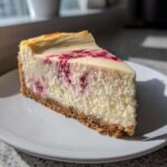 A rich slice of white chocolate cheesecake with a raspberry swirl on a white plate.