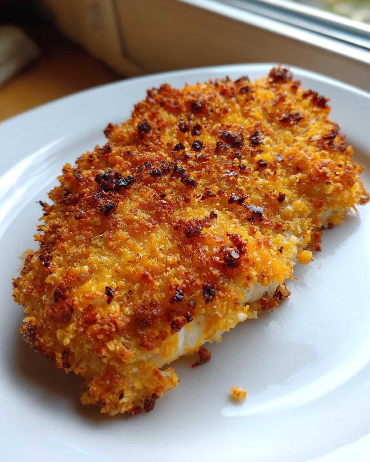 Close-up of a golden brown, baked crispy cheddar chicken fillet on a white plate.