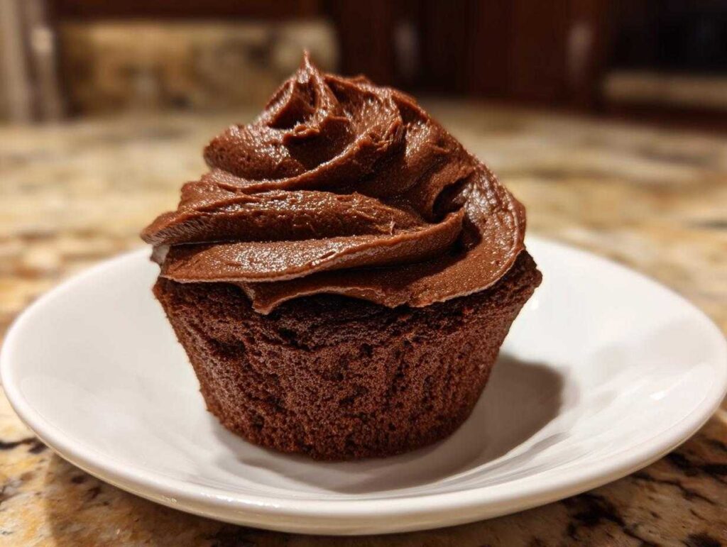 A single chocolate cupcake topped with a generous swirl of rich chocolate frosting on a small white plate.