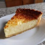 A close-up of a thick slice of creamy custard pie with a golden-brown, slightly textured top, resting on a white plate.