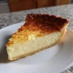 A close-up of a thick slice of creamy custard pie with a golden-brown, slightly textured top, resting on a white plate.