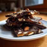 Stack of dark chocolate almond toffee pieces showing gooey caramel layer on a white plate.