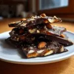 Stack of dark chocolate almond toffee pieces showing gooey caramel layer on a white plate.