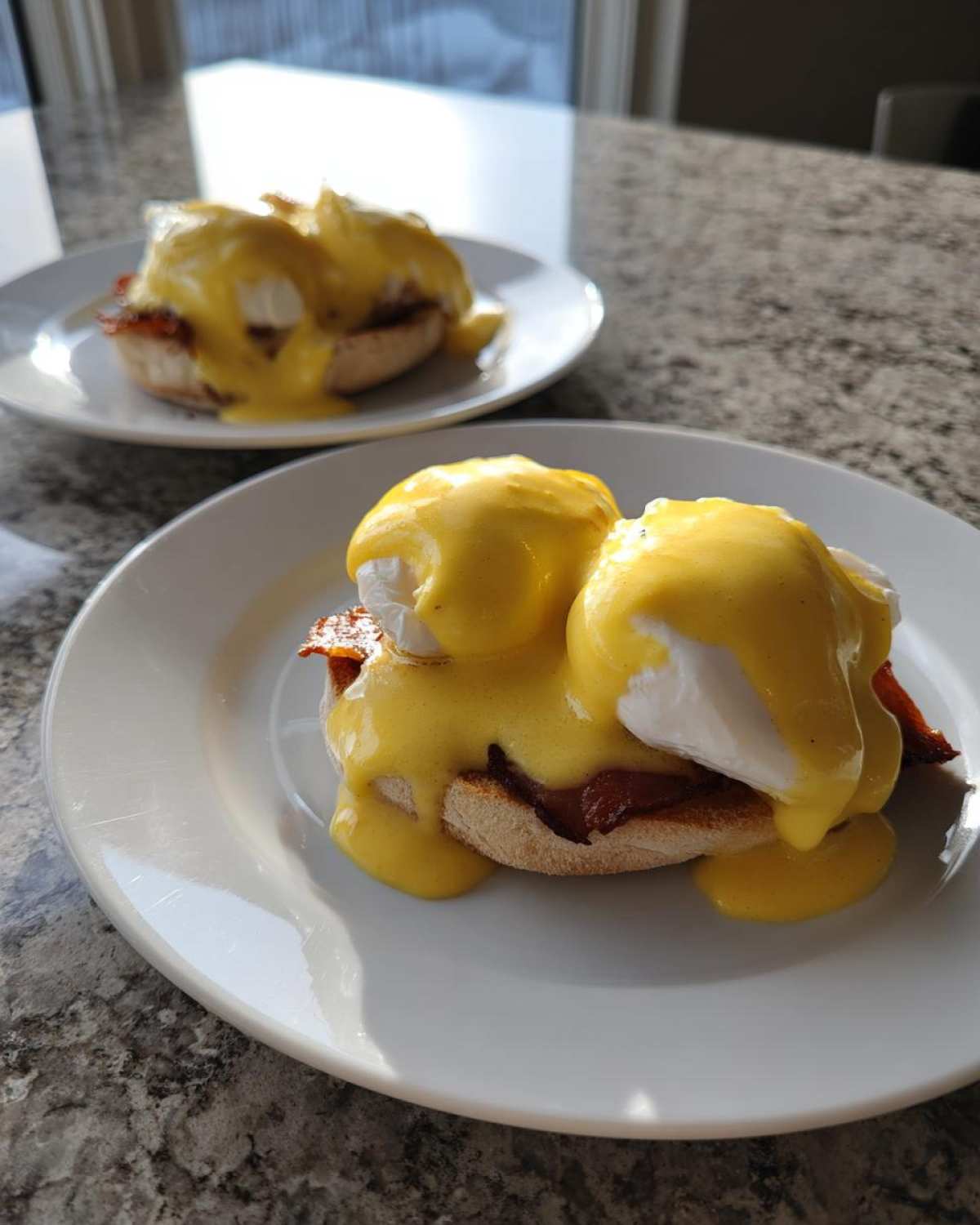 Close-up of two servings of eggs benedict smothered in hollandaise sauce on white plates.