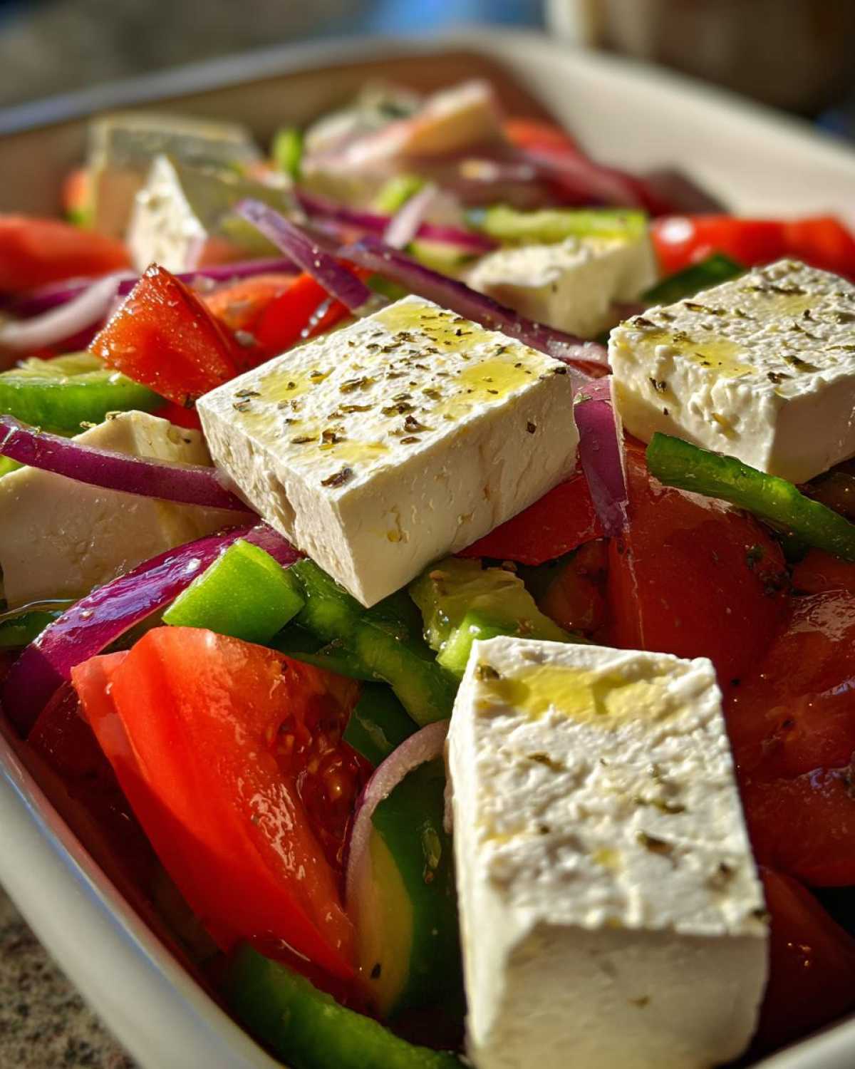 Close-up of thick feta cheese blocks drizzled with olive oil in a vibrant greek salad with tomatoes and red onion.