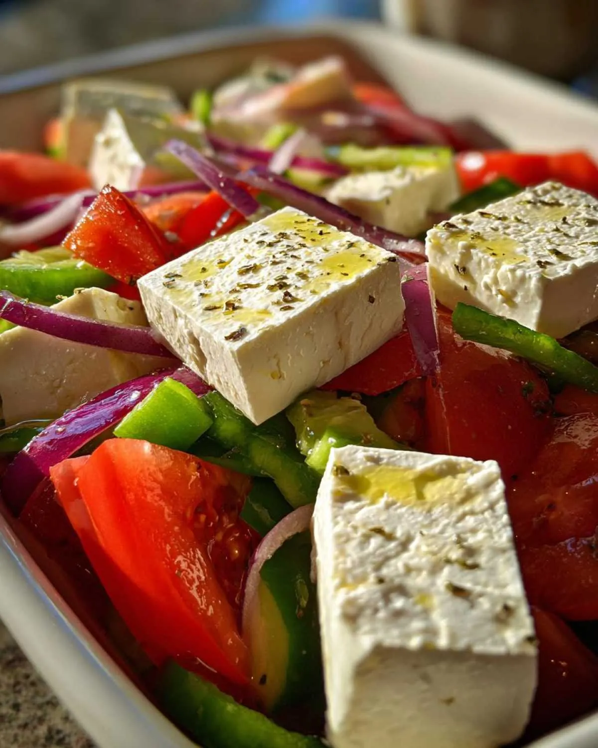 Close-up of thick feta cheese blocks drizzled with olive oil in a vibrant greek salad with tomatoes and red onion.
