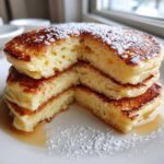 A stack of fluffy greek yogurt pancakes cut to show the airy interior, dusted with powdered sugar and drizzled with syrup.