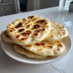 A stack of freshly cooked, fluffy naan bread with golden-brown charred spots resting on a white plate.