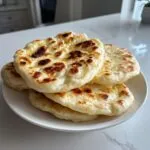 A stack of freshly cooked, fluffy naan bread with golden-brown charred spots resting on a white plate.