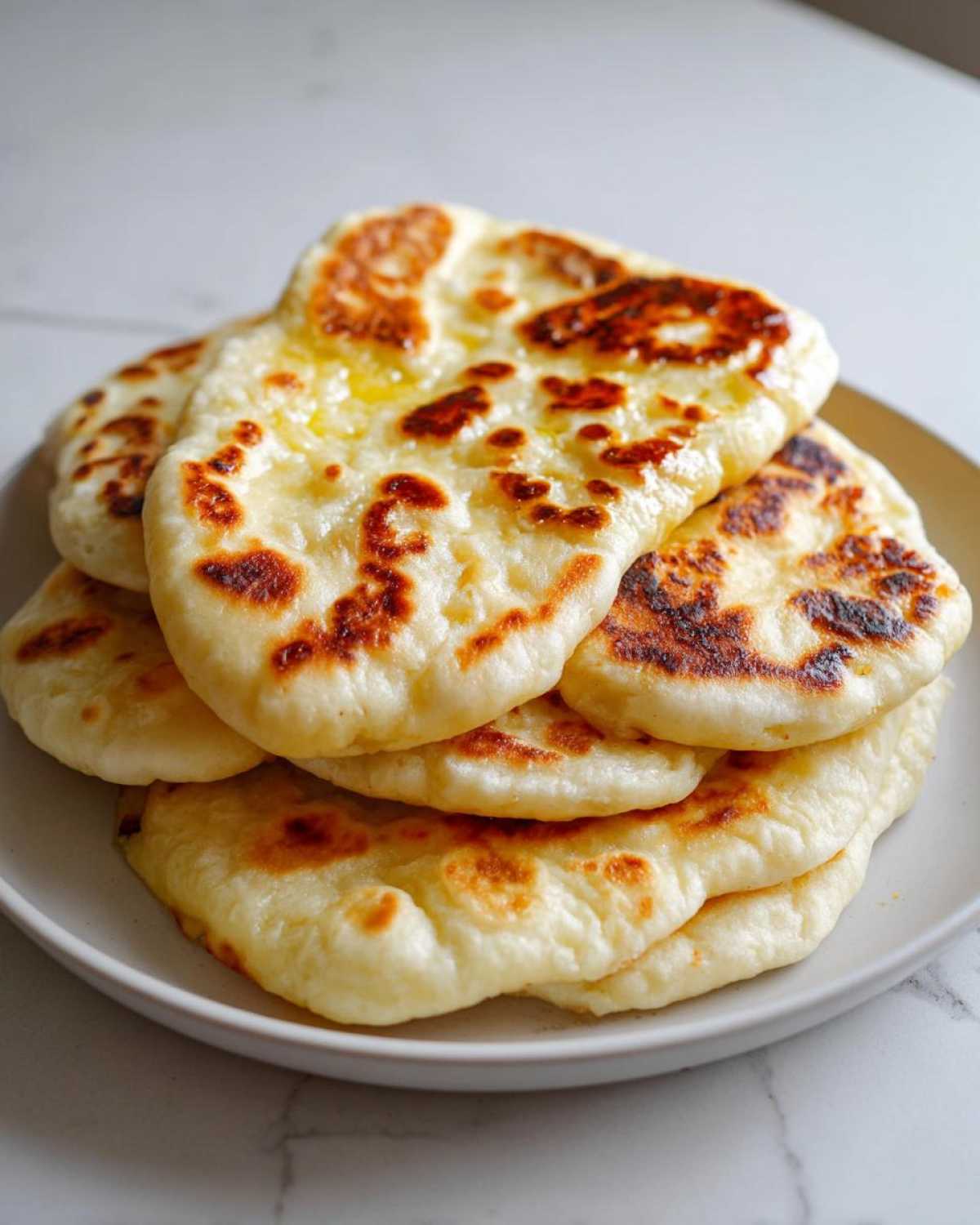 A stack of freshly made, soft naan bread pieces glistening with melted butter, featuring charred spots.