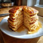 A tall stack of fluffy almond flour pancakes cut in half, drizzled generously with maple syrup on a white plate.
