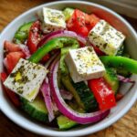 Close-up of a vibrant greek salad featuring large cubes of feta cheese drizzled with oil and herbs, mixed with cucumber, tomato, and red onion.