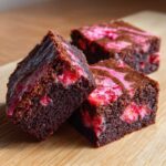 Three fudgy chocolate strawberry brownies with visible pink swirls and fruit pieces on a wooden surface.