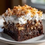 A moist slice of german chocolate poke cake topped with whipped cream and toasted coconut flakes.