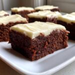 A close-up of a rich, dark gingerbread brownies square topped with thick, creamy white frosting.