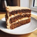 A tall slice of three-layer gingerbread latte cake with thick, spiced frosting layers, sitting on a white plate.