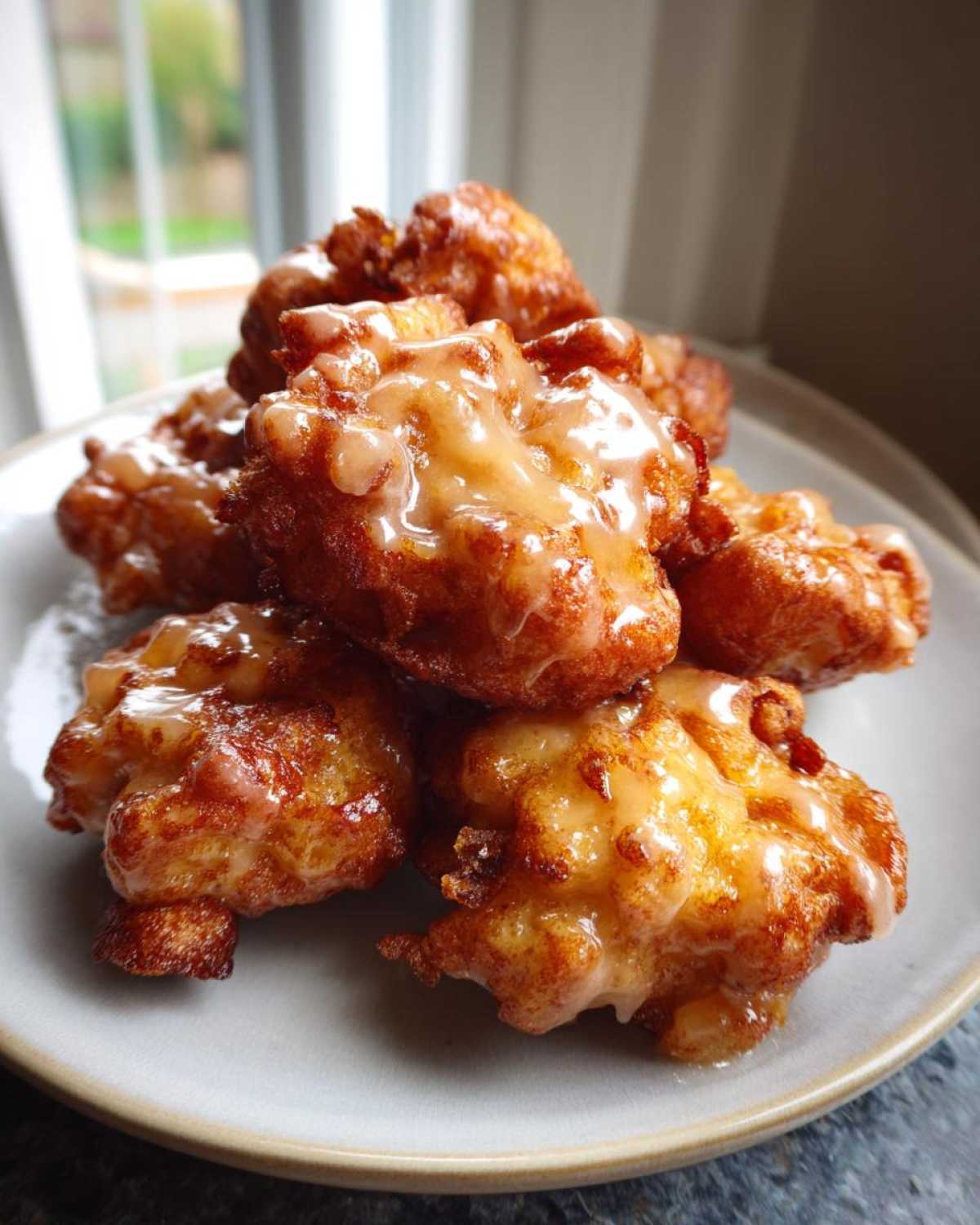 A stack of golden brown, glazed apple fritter bites piled high on a light-colored plate.
